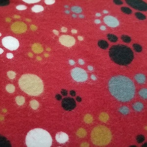 May include: Red fabric with a repeating pattern of paw prints in white, black, yellow, and light blue. The paw prints are scattered across the surface, creating a playful and whimsical design. The fabric has a soft, textured appearance.