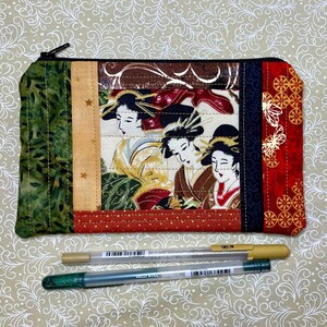 May include: A rectangular, quilted fabric pouch with a zipper closure. The pouch features a patchwork design with green, tan, and red panels. The central panel displays a print of three geishas. Two pens are visible below the pouch.