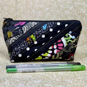 May include: A quilted cosmetic bag with a black zipper, featuring a patchwork design of various patterned fabrics in black, white, and colorful accents. Two gel pens with green and gray caps are in front of the bag.