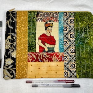 May include: A quilted zippered pouch with a colorful patchwork design featuring a portrait of Frida Kahlo. The pouch is made of various fabrics with different patterns, including floral, geometric, and paisley. The pouch is a great way to store small items like pens, pencils, and makeup.