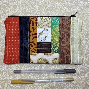May include: A quilted zippered pouch with a patchwork design featuring various fabrics in shades of brown, green, red, and black. The pouch has a small embroidered image of a cat wearing glasses.