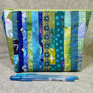 May include: A green zippered pouch with a patchwork design of blue, green, and yellow fabric strips. The pouch is lying on a patterned surface with two blue gel pens.