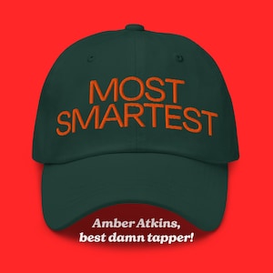 May include: A dark green baseball cap featuring the embroidered orange text MOST SMARTEST on the front. Below the cap, white text reads Amber Atkins, best damn tapper! The cap has a structured crown, a curved brim, and a button on top.