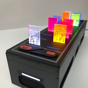 May include: A black box with a row of colorful translucent cards with numbers on them. The cards are lit up with different colors. The box has a red button and a white light.