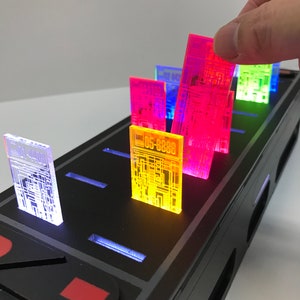 May include: A black electronic device with a row of colorful translucent circuit board-like chips. The chips are lit from below, creating a glowing effect. The device has a red button and a black surface.