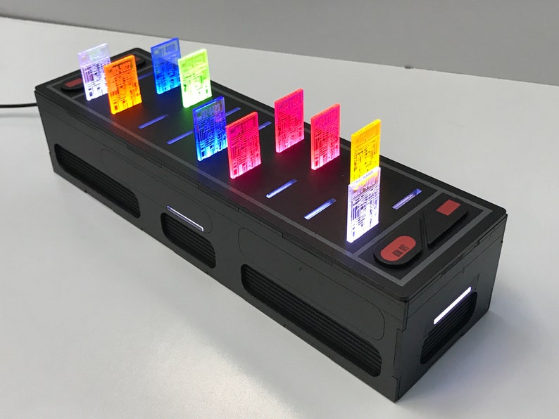 May include: A black box with a row of illuminated translucent circuit boards in various colors, including red, orange, yellow, green, blue, and purple. The box has a red button labeled "SYS".