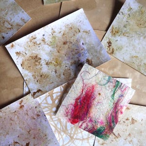 May include: A collection of rectangular handmade paper sheets featuring textured surfaces with embedded organic brown plant fibres and one square piece accented with vibrant red, pink, and green thread fibres layered across the off-white paper base.