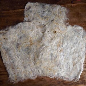 May include: A handmade sheet of textured paper with a rough, uneven surface. The paper is mainly off-white with subtle brown and grey tones, and a visible fibre structure. The sheet is laid out on a wooden surface.