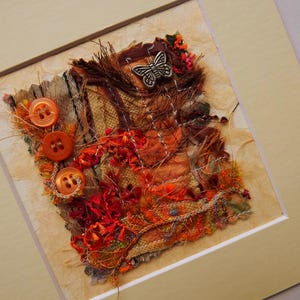 May include: Textile artwork featuring a collage of textured fabrics in shades of brown, orange, and red. Includes three orange buttons and a silver butterfly embellishment. The artwork is framed in a cream-coloured mount.