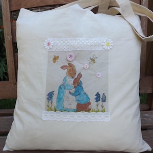 May include: A cream-coloured canvas tote bag featuring a central rectangular patch with a printed illustration of two rabbits in blue jackets. The patch is framed by white lace trim, small fabric flowers, and three light pink buttons.