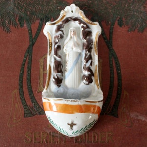 Könnte beinhalten: Ein weißer Porzellan-Wandhalter mit goldener Verzierung und einer Darstellung der Jungfrau Maria in einem blauen Gewand. Der Halter hat ein orangefarbenes Band und grüne Blätter am unteren Rand.