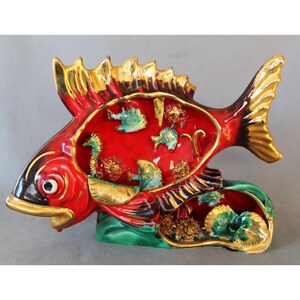 Könnte beinhalten: Dekorative Keramikfigur in Fischform, hauptsächlich rot mit goldenen Akzenten. Der Fisch hat ein offenes Inneres mit Meeresbewohnern und steht auf einem grünen Sockel. Die Flossen und der Schwanz sind ebenfalls golden detailliert.