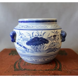 Pode incluir: Um vaso de cerâmica branca com motivos florais e de folhas pintados à mão em azul. O vaso apresenta um corpo arredondado, uma abertura larga e duas pequenas alças azuis nas laterais. O design inclui faixas azuis horizontais nas bordas.