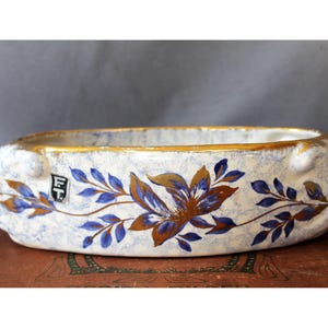 May include: An oval ceramic bowl with a white and blue mottled finish, featuring a gold-painted rim and a floral motif in blue and metallic gold. A small rectangular label with the letters FT is visible on the side of the bowl.