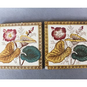Può includere: Due piastrelle quadrate in ceramica con motivi floreali. Ogni piastrella presenta un fiore centrale nei toni del rosso e del rosa, circondato da foglie gialle e verdi. Le piastrelle hanno un bordo decorativo.