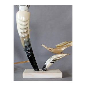Op de afbeelding: Een unieke tafellamp met een hoornvormige basis, die overloopt van crème naar zwart. Twee decoratieve vogelsculpturen zijn aan de basis bevestigd. De lamp staat op een rechthoekige, gebroken witte basis. De lamp heeft een standaard lampfitting.