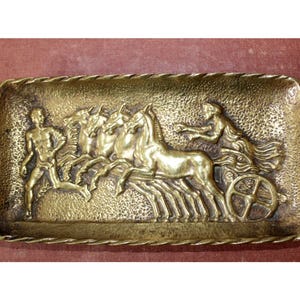Może przedstawiać: Prostokątna mosiężna taca z reliefem przedstawiającym wyścig rydwanów. Taca ma teksturowaną powierzchnię i plecioną obwódkę. Scena obejmuje biegacza, konie, woźnicę i koło rydwanu.