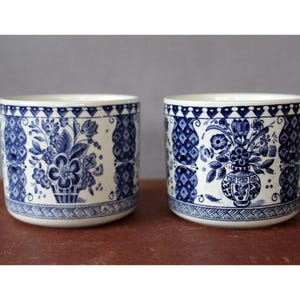 Puede incluir: Dos recipientes de cerámica a juego con un diseño floral azul y blanco. Los recipientes presentan un patrón repetitivo de flores y formas geométricas. Los bordes y las bases tienen un borde detallado. Los recipientes son cilíndricos y se asientan sobre una superficie marrón.