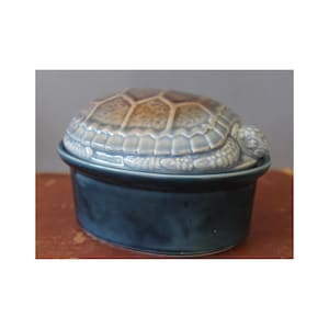 Könnte beinhalten: Eine keramische Schmuckschatulle in Schildkrötenform. Der Deckel ist ein detaillierter Schildkrötenpanzer in Braun- und Blautönen, wobei Kopf und Flossen der Schildkröte sichtbar sind. Der Sockel ist ein fester, dunkelblauer, oval geformter Behälter.