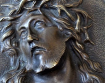 Könnte beinhalten: Bronze-Reliefskulptur, die einen Mann mit Bart und Dornenkrone darstellt. Die Skulptur ist auf einer kreuzförmigen Platte mit strukturiertem Hintergrund und goldfarbenem Rand montiert. Das Gesicht des Mannes zeigt einen ruhigen Ausdruck.
