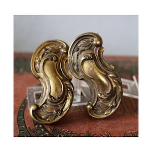 Könnte beinhalten: Zwei antike Schubladengriffe aus Messing mit einem verzierten, geschwungenen Design. Die Griffe weisen ein detailliertes, symmetrisches Muster aus Wirbellinien und blattartigen Formen auf. Das Messing hat eine warme, gealterte Patina.