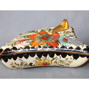 Peut inclure: Boîte à bijoux décorative peinte à la main avec un motif floral. La boîte crème a des bords festonnés noirs et une poignée dorée. Le couvercle présente des fleurs orange, bleues et jaunes avec des feuilles vertes. Mesure environ 15 cm de long.