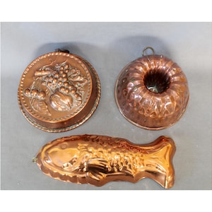 Op de afbeelding: Drie koperen bakvormen, waaronder een ronde vorm met reliëf van fruit, een ronde geribbelde tulbandvorm met een ophanglus en een visvormige bakvorm. Elk stuk heeft een metallic koperen afwerking en gedetailleerde reliëfpatronen voor het bakken.