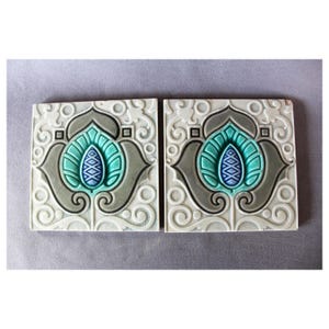 May include: Two ceramic tiles featuring a decorative floral design. The tiles are white with a grey and blue floral pattern. The flowers are stylised and have a blue centre.
