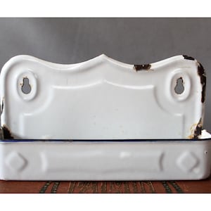May include: White enamel wall-mounted shelf with a decorative top and a rectangular base. The shelf has two keyhole-shaped mounting holes and a dark blue trim. The enamel shows signs of wear, adding to its vintage appeal.