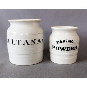 May include: Two white ceramic kitchen canisters. One reads "SULTANAS" in black lettering, the other reads "BAKING POWDER". Both have a cylindrical shape with a slightly flared rim, and horizontal ridges.