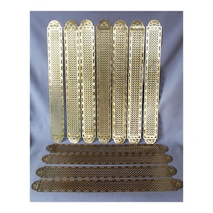 May include: A collection of brass-colored, decorative metal strips. The longer strips have an elongated oval top and intricate cut-out patterns. Shorter strips with similar designs are below. The metal has a polished finish.