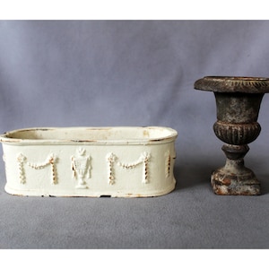 May include: An oval-shaped off-white planter with embossed garland and urn motifs alongside a small, dark, weathered metal pedestal urn featuring a wide, fluted rim and a square base. Both items exhibit signs of age and textured, worn surfaces.
