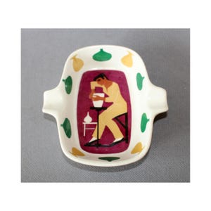 Op de afbeelding: Een vintage keramische asbak met een witte basis, met een centraal rechthoekig ontwerp met een figuur in geel en zwart. Het ontwerp is geplaatst tegen een bordeauxrode achtergrond, met groene en gele vormen rond de rand.