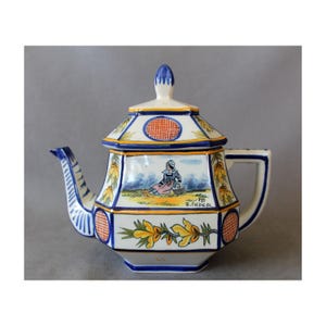 Può includere: Una teiera in ceramica bianca con un motivo floreale blu e giallo. La teiera ha un corpo ottagonale e un coperchio a cupola. La teiera è decorata con una scena di una donna seduta in un campo. La teiera è firmata "HB RIMPER".