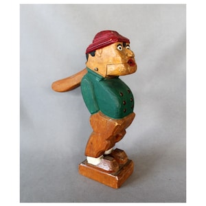 May include: A vintage wooden nutcracker shaped like a man. It features a red hat, green jacket, and brown trousers. The nutcracker has a lever arm and is mounted on a wooden base. The figure is approximately 20 cm tall.