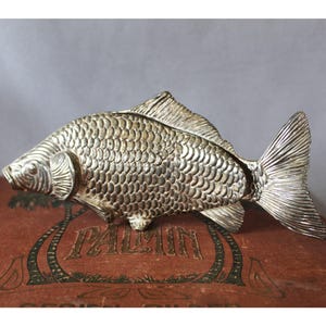 Pode incluir: Uma estatueta de peixe em metal prateado com padrões de escamas detalhados, uma cabeça definida e uma grande barbatana caudal texturizada. O peixe está posicionado horizontalmente, exibindo um acabamento metálico com reflexos e sombras.