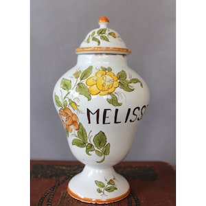 May include: A white ceramic apothecary jar with a matching lid, featuring hand-painted yellow and orange floral motifs and green leaves. The word MELISSE is written in dark brown lettering on the front. The jar has a pedestal base and orange trim.