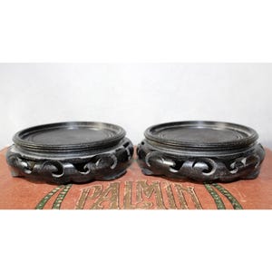 Könnte beinhalten: Zwei dunkelgraue, geschnitzte Holzständer mit kreisförmiger Oberseite und dekorativem, durchbrochenem Sockel. Die Ständer werden auf einer rotbraunen Oberfläche mit verblasstem Text gezeigt.