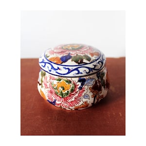 Puede incluir: Una caja redonda de cerámica con tapa, decorada con un patrón floral en tonos de rosa, azul, verde, amarillo y marrón. La caja tiene un fondo blanco y un borde azul y blanco.