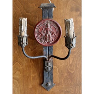 May include: A dark metal wall sconce featuring two candle holders with weathered white wax remnants. A central circular red medallion displays a relief of a seated figure holding a staff, positioned above a small metal fleur-de-lis emblem.