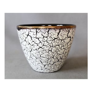 Puede incluir: Un cuenco de cerámica blanca con un patrón de esmalte craquelado y un borde dorado. El cuenco tiene una abertura ancha y se estrecha hacia la base. El interior es negro.