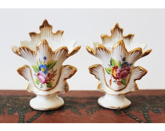 Könnte beinhalten: Zwei weiße Porzellanvasen mit goldenen Akzenten und Blumendesigns. Die Vasen sind mit handbemalten Blumen in Rosa-, Blau- und Gelbtönen verziert.