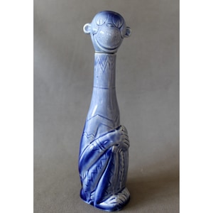 Könnte beinhalten: Eine blaue Keramikflasche mit einem affenförmigen Verschluss. Die Flasche hat einen langen Hals und ein detailliertes Design eines Affen, der eine Decke hält. Das Gesicht des Affen lächelt, und die Flasche ist hellblau.