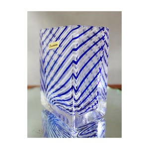 May include: A clear glass vase with a square shape and a pattern of blue and white diagonal stripes. A small label with the word "Prudence" is visible on the side of the vase. The vase is reflected on a glass surface.