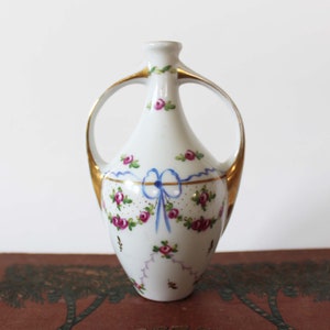 May include: A small white porcelain vase with gold accents and floral designs. The vase has two handles and a delicate blue ribbon with white polka dots.