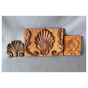 May include: Three wooden carvings of seashells. The largest carving is a rectangular piece with a seashell in the centre and ornate scrollwork on either side. The other two carvings are smaller and feature a single seashell.