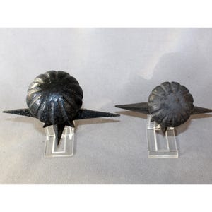 May include: Two black metal decorative objects with a starburst design. Each object has a round, textured top and pointed extensions. They are displayed on clear plastic stands against a neutral background.