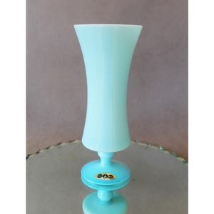 May include: A light blue glass vase with a flared top and a round base. The vase has a slender neck and a small, circular base. A small, oval sticker is on the base. The vase is sitting on a reflective surface.