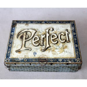 May include: A rectangular metal tin box with a weathered white and blue finish. The lid features the embossed word Perfect in large lettering, surrounded by floral illustrations and a small arrow symbol. The sides have a blue and white pattern.