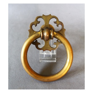Op de afbeelding: Een messing ladeknop met een ronde handgreep en sierlijke achterplaat. De knop heeft een gouden kleur en is waarschijnlijk gemaakt van massief messing. Het ontwerp heeft een ringhandvat en decoratieve versieringen.